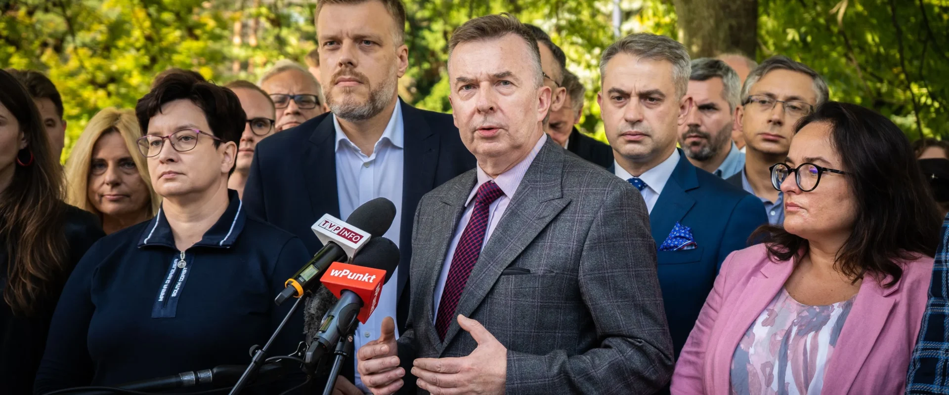 Na zdjęciu widoczna jest grupa osób, w tym mężczyzna mówiący do mikrofonów, znajdujący się w otoczeniu innych ludzi, którzy słuchają jego wypowiedzi.