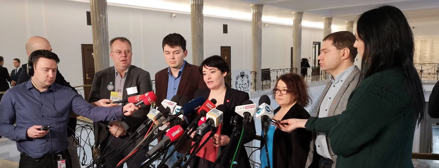 Grupa polityków i dziennikarzy udziela konferencji prasowej, stojąc przed licznymi mikrofonami w jasnym wnętrzu z kolumnami.