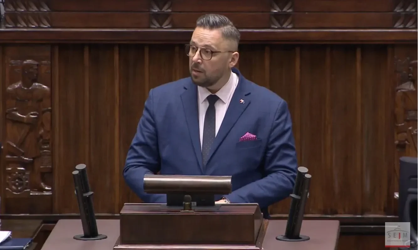 Mężczyzna w granatowym garniturze z różową poszetką stoi przy mównicy w sali plenarnej z drewnianymi panelami w tle i wygłasza przemówienie.