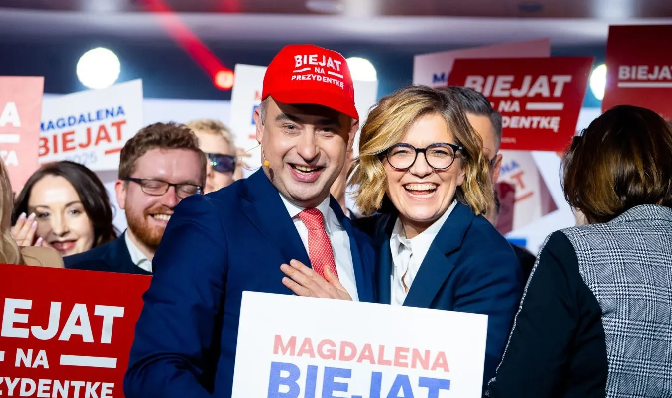 Grupa uśmiechniętych uczestników wiecu Lewicy trzyma plakaty z hasłami wyborczymi, a mężczyzna w czerwonej czapce z napisem „BIEJAT NA PREZYDENTKĘ” stoi obok kobiety.