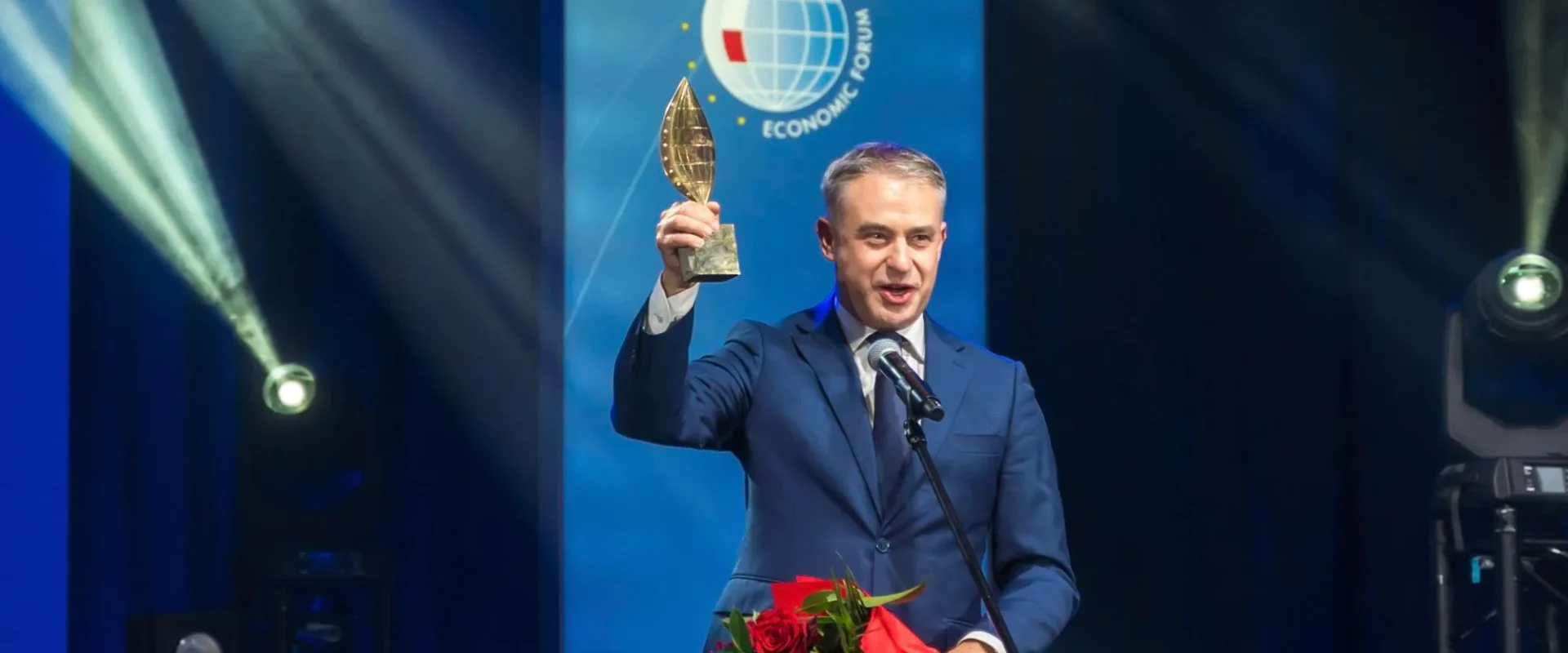 Mężczyzna w niebieskim garniturze stoi na scenie, unosi nagrodę w kształcie liścia i trzyma bukiet czerwonych róż podczas Forum Ekonomicznego w Karpaczu.