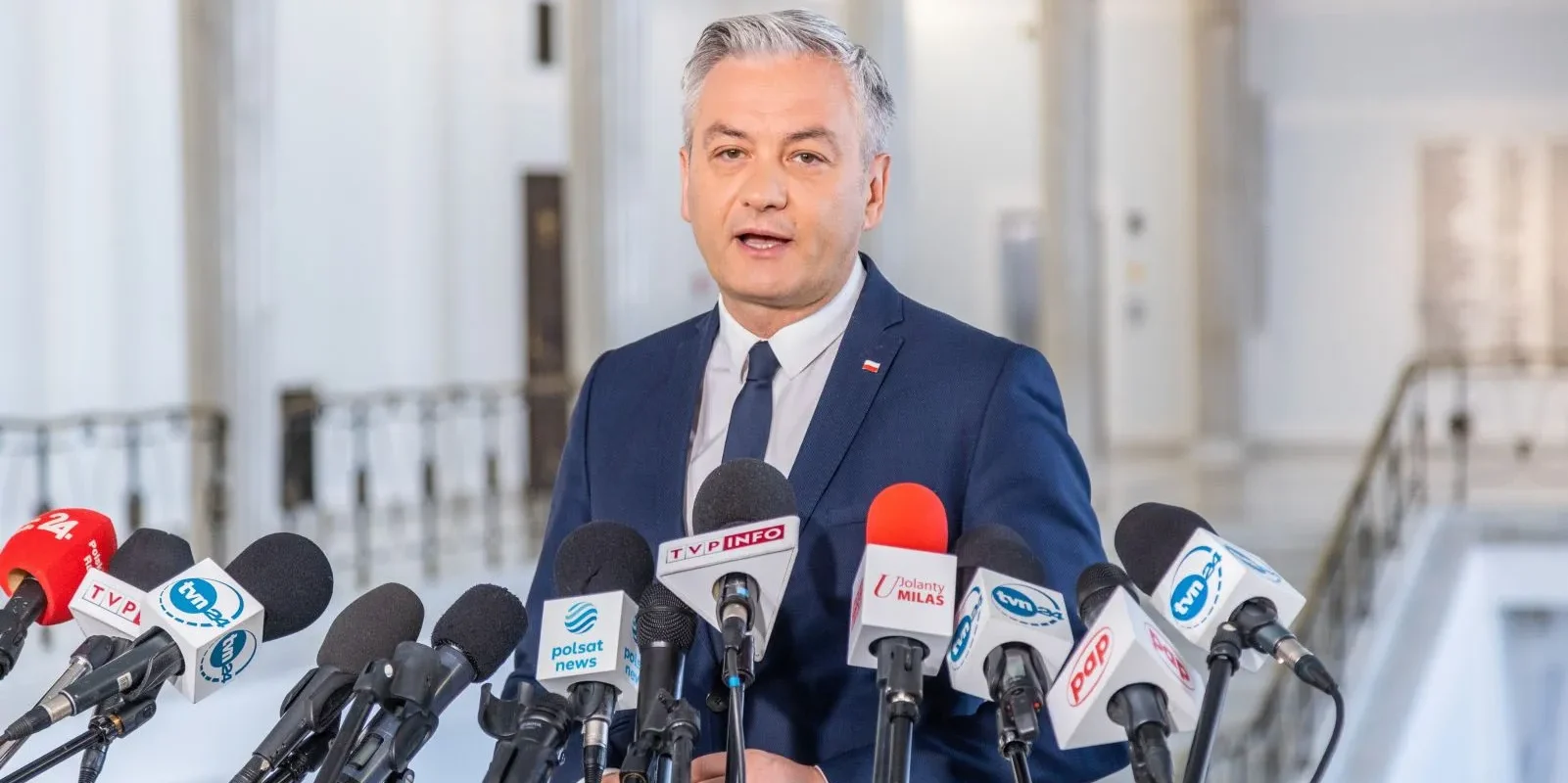 Na zdjęciu widać mężczyznę w garniturze, stojącego przed mikrofonami na konferencji prasowej w jasnym wnętrzu.
