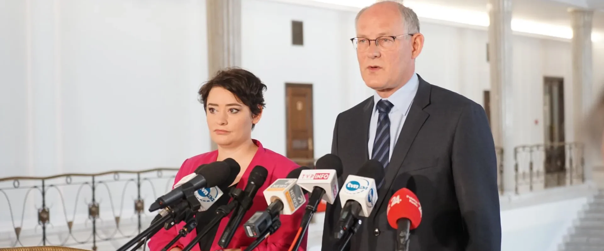 Dwie osoby stoją przy zestawie mikrofonów podczas konferencji prasowej w jasnym holu z kolumnami.