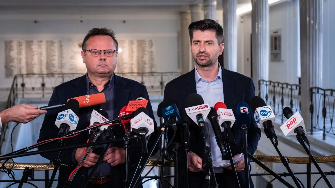 Na zdjęciu dwóch mężczyzn stoi obok siebie przed mikrofonami, jeden z nich mówi, podczas gdy drugi słucha, w tle widoczna jest elegancka architektura.