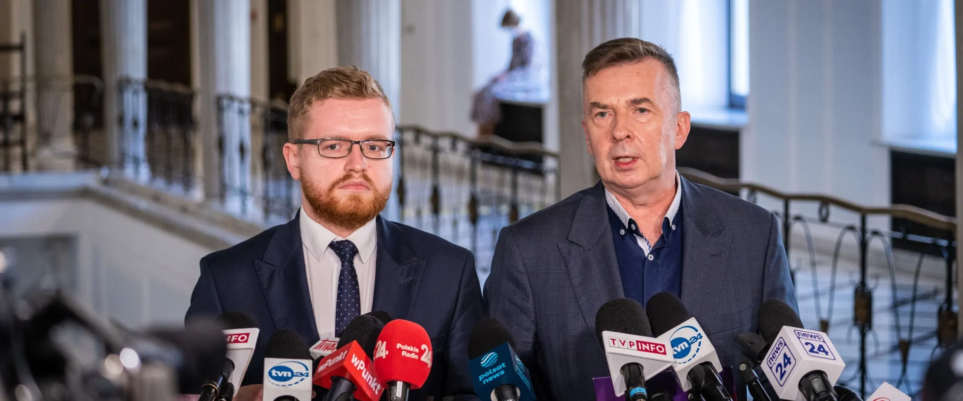 Dwóch mężczyzn w garniturach udziela konferencji prasowej, stojąc przed zestawem mikrofonów.