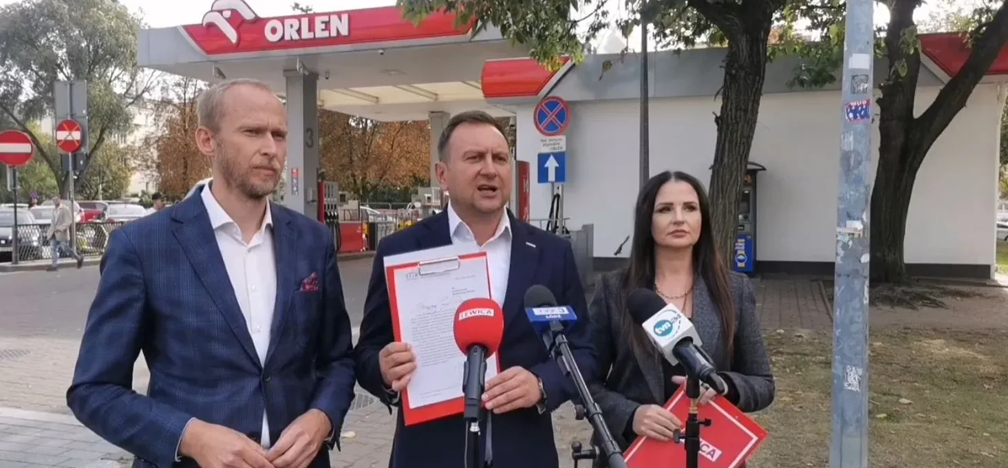 Trzech uczestników konferencji prasowej stoi przed stacją Orlen, trzyma dokumenty i przemawia do mikrofonów.