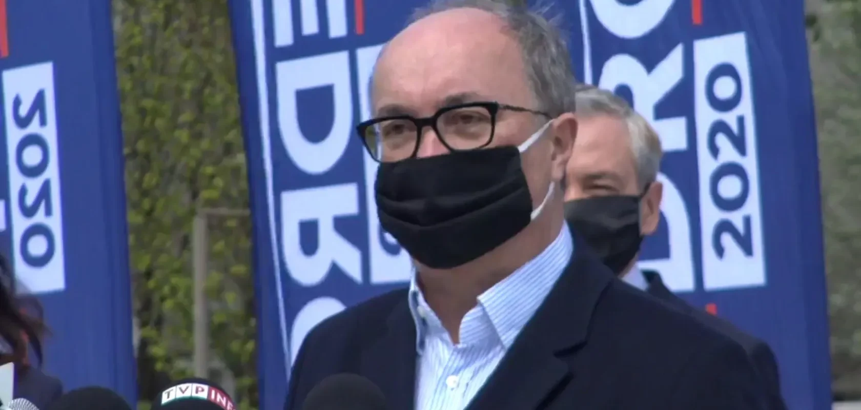 Mężczyzna w maseczce i okularach przemawia do mediów podczas konferencji prasowej, stojąc przed niebieskimi flagami.