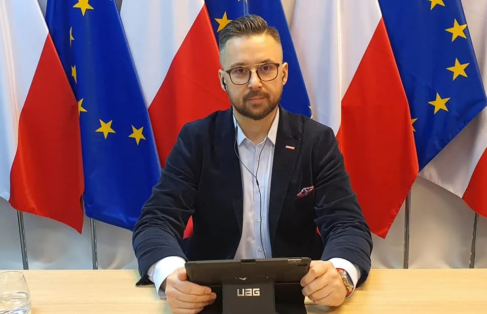 Mężczyzna w garniturze i okularach siedzi za biurkiem, trzyma tablet, a w tle stoją flagi Polski i Unii Europejskiej.