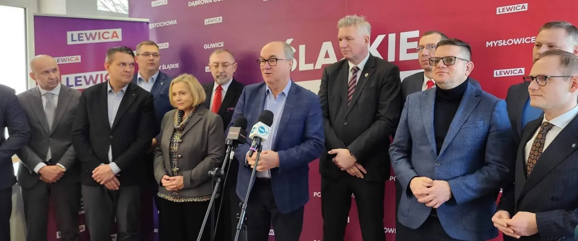 Na zdjęciu widać grupę mężczyzn i kobiet stojących razem przed bannerem z napisem "LEWICA", z jednym z mężczyzn mówiącym do mikrofonów.