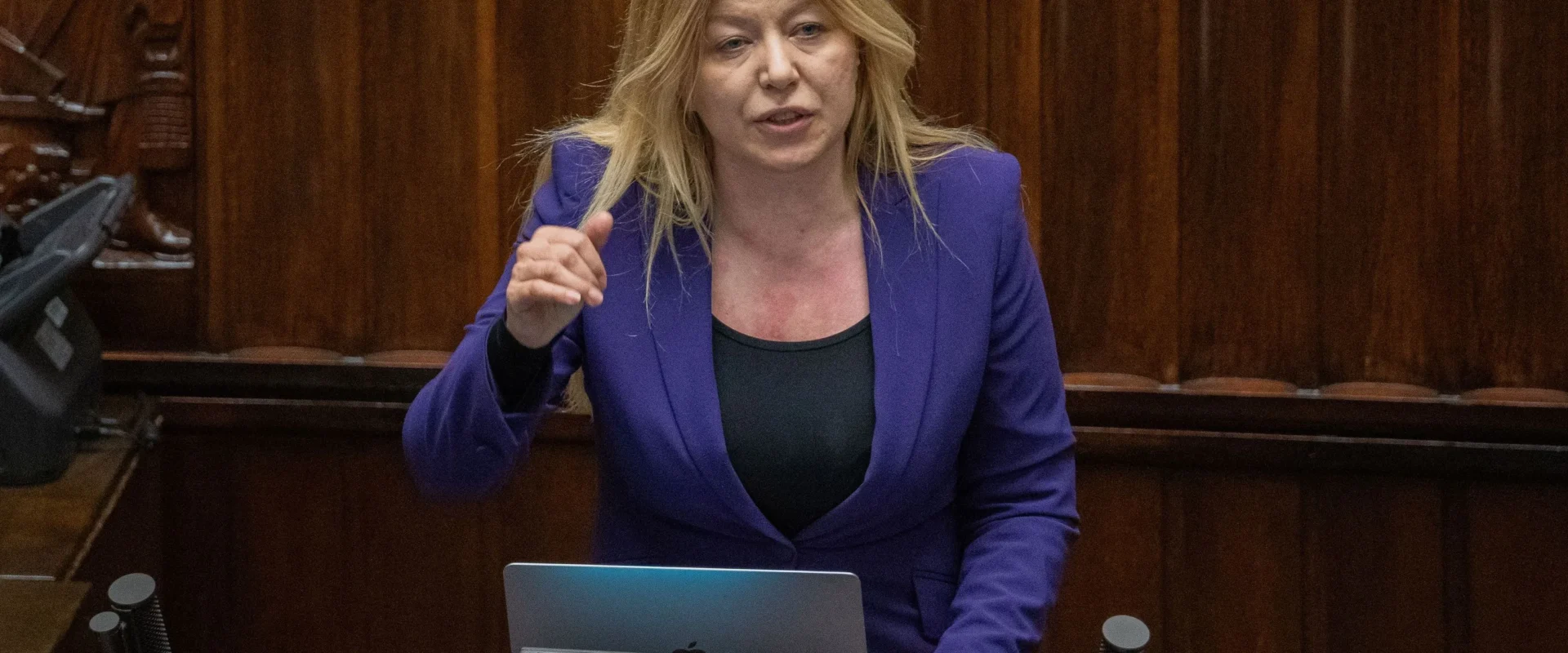 Kobieta blond włosów w fioletowej marynarce przemawia z laptopem na mównicy w sali plenarnej.