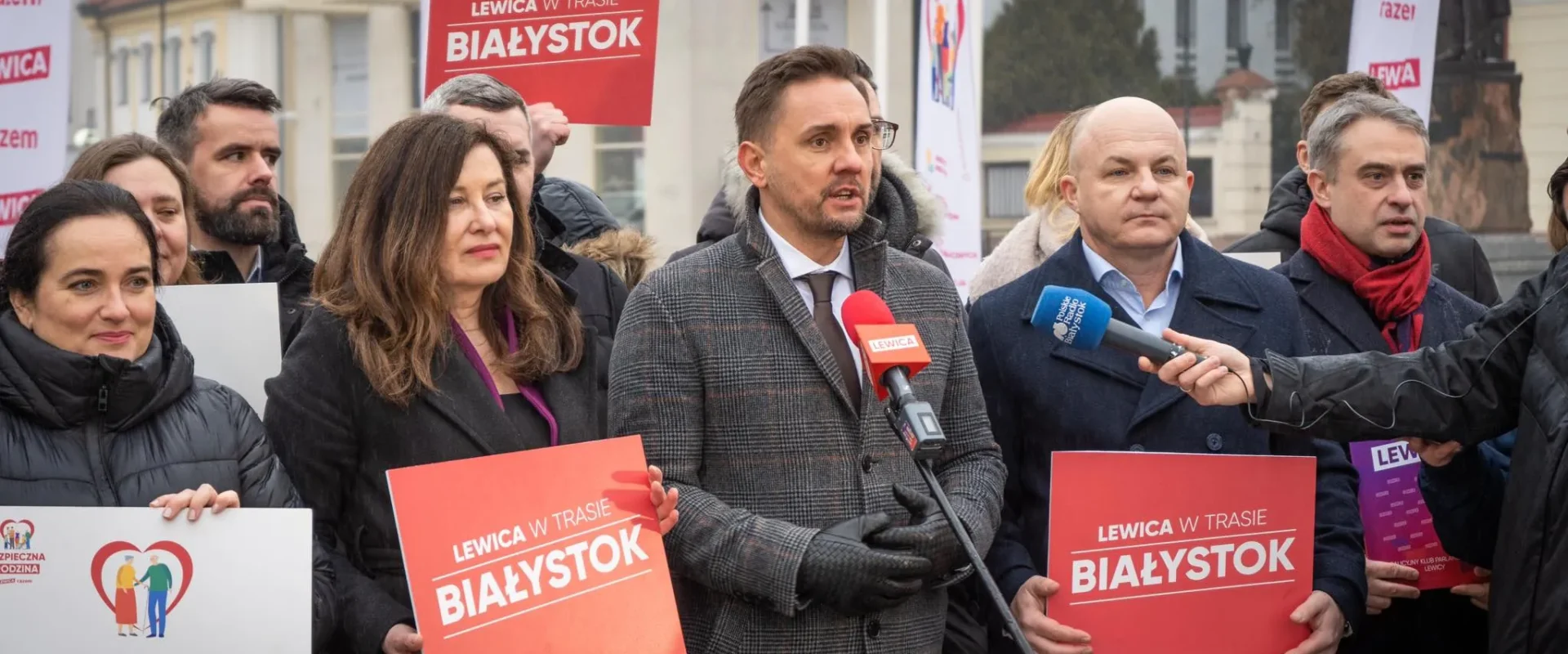Na zdjęciu grupa osób z partii Lewica prowadzi konferencję prasową w Białymstoku, trzymając transparenty i plakaty dotyczące aktu politycznego.