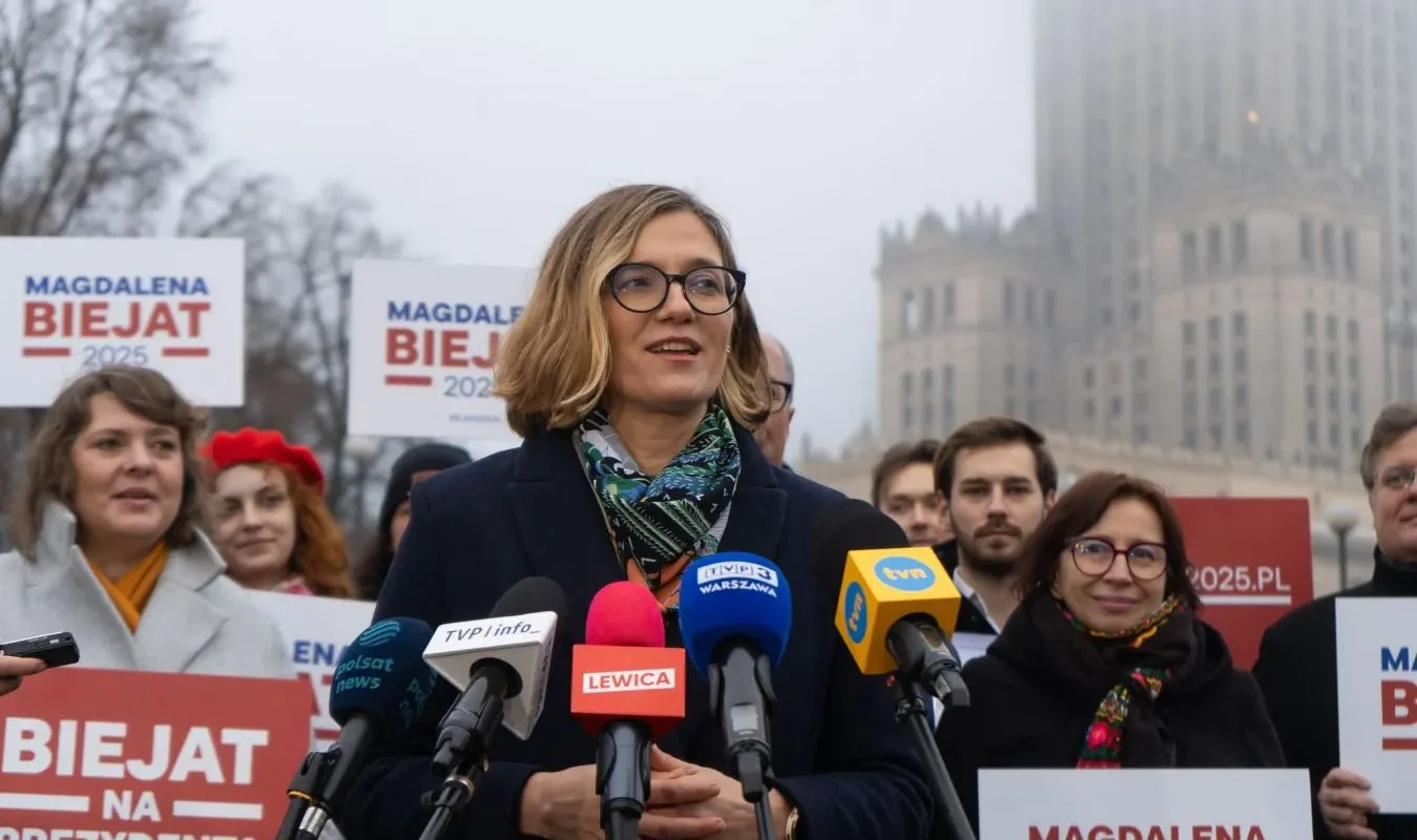 Kobieta w ciemnym płaszczu i okularach przemawia na konferencji prasowej przed zestawem mikrofonów, otoczona innymi osobami i plakatami z napisem „BIEJAT” na tle miejskiego krajobrazu.