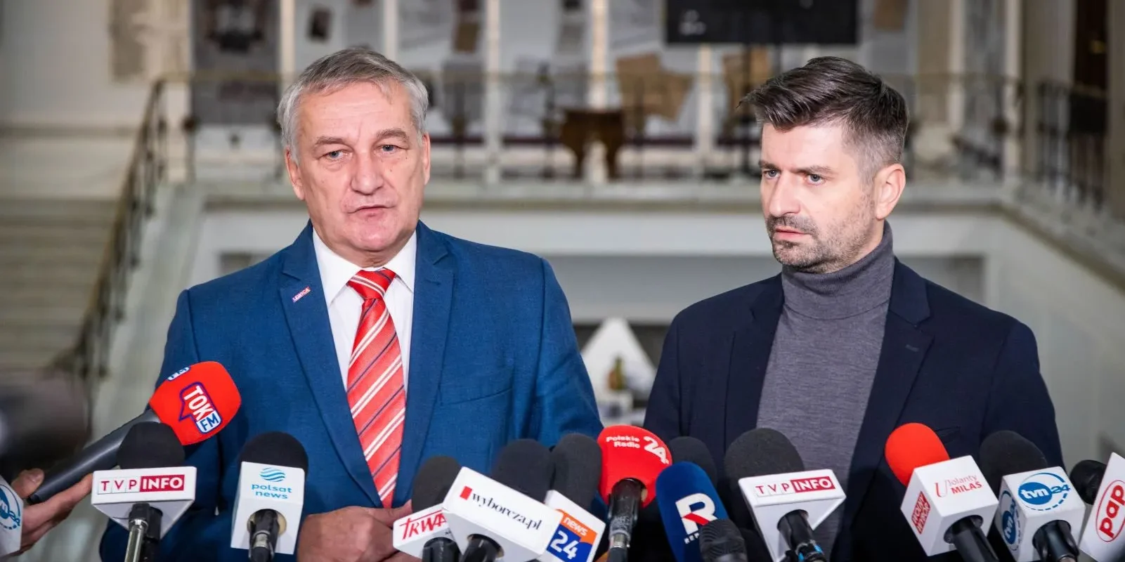 Na zdjęciu widać dwóch mężczyzn stojących przed mikrofonami i kamerami w obszernym wnętrzu, prawdopodobnie podczas konferencji prasowej.
