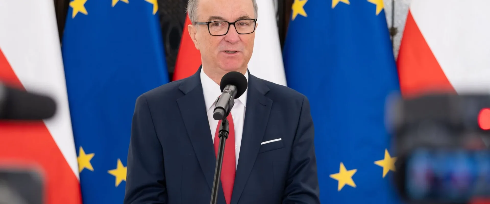 Mężczyzna w ciemnym garniturze z czerwonym krawatem przemawia do mikrofonu na tle flag Unii Europejskiej i Polski.