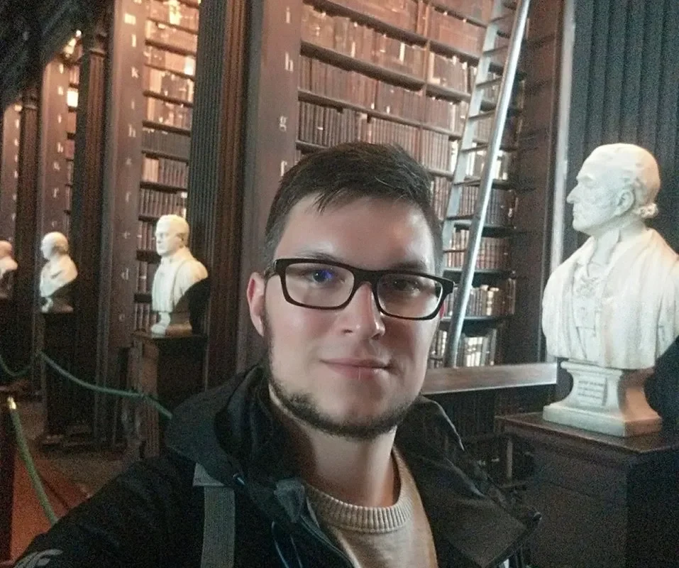 Mężczyzna w okularach robi selfie w zabytkowej bibliotece, stojąc przed wysokimi regałami z książkami i białymi rzeźbami-bustami.