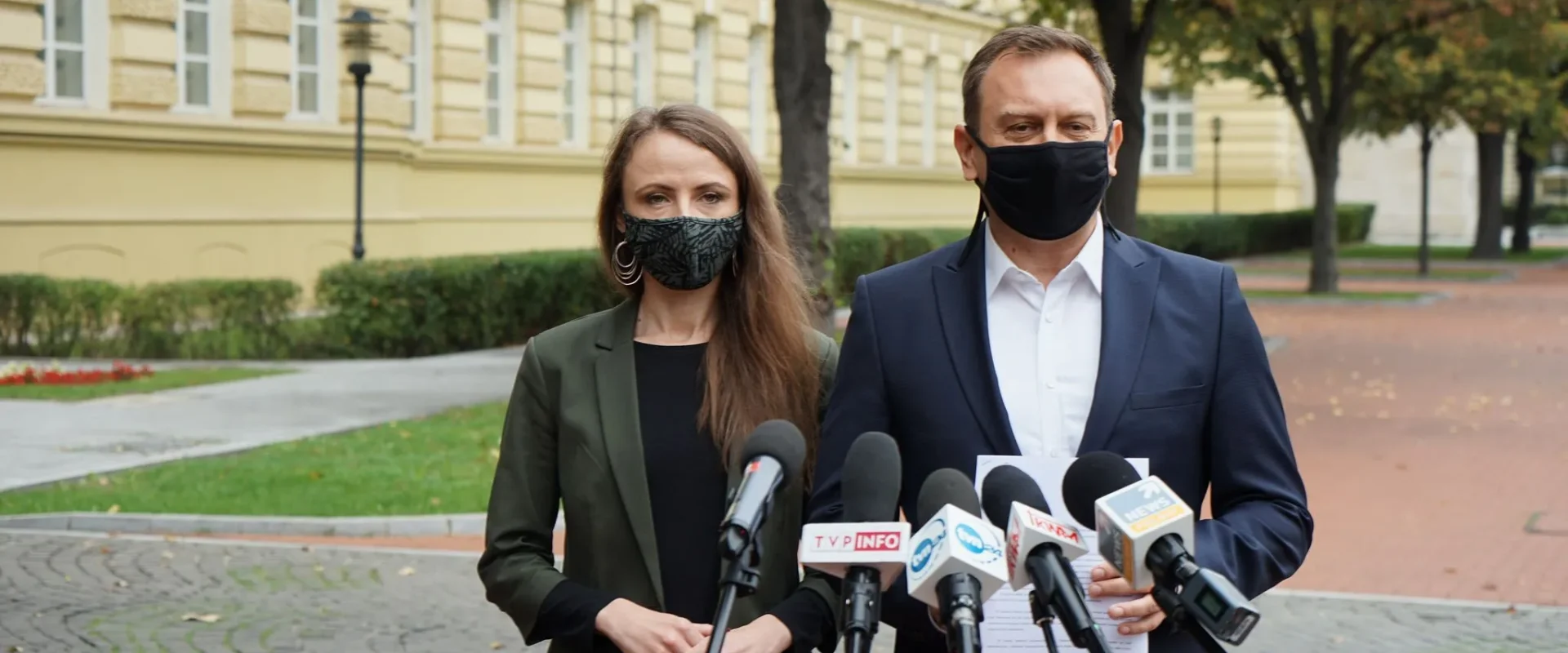 Mężczyzna i kobieta w maseczkach stoją na zewnątrz przed zestawem mikrofonów podczas konferencji prasowej, na tle żółtej elewacji budynku.