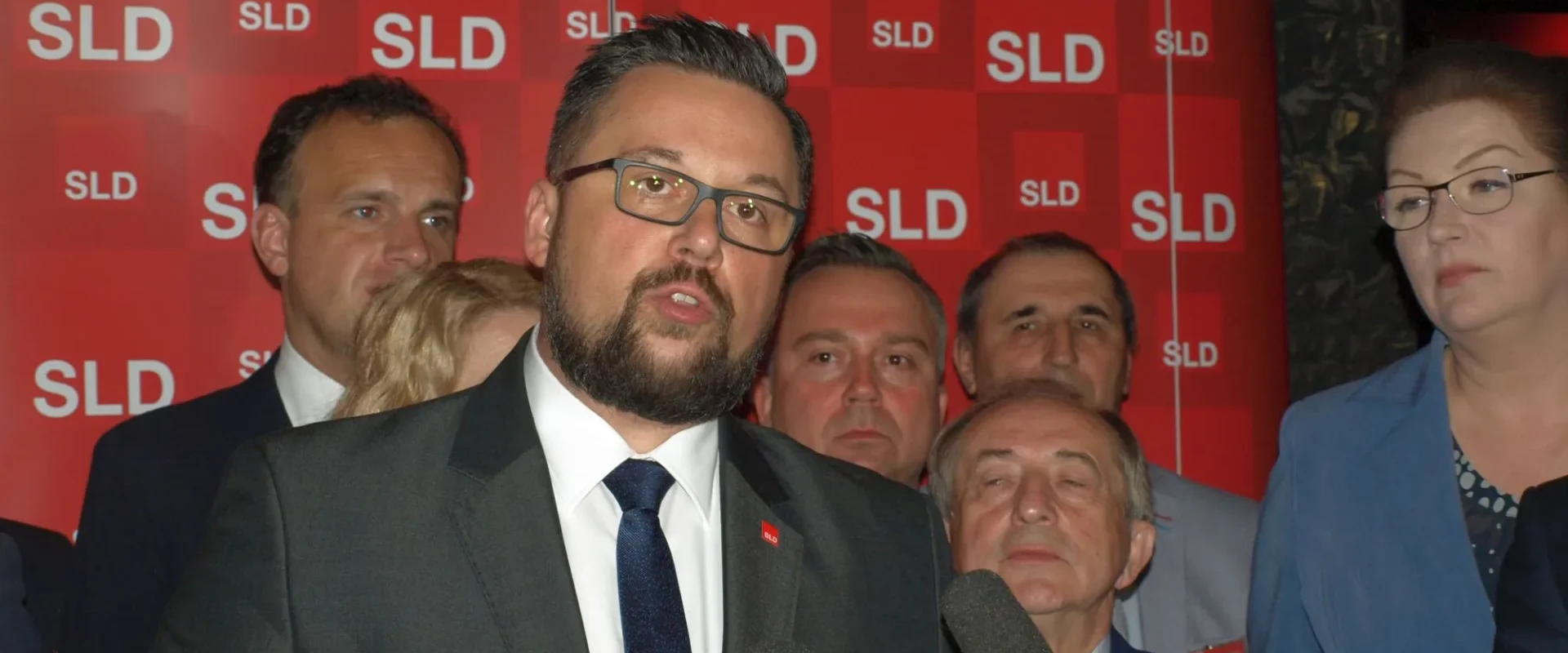Mężczyzna w ciemnym garniturze z brodą i okularami przemawia do mikrofonów podczas wystąpienia na tle czerwonego baneru z logo SLD.