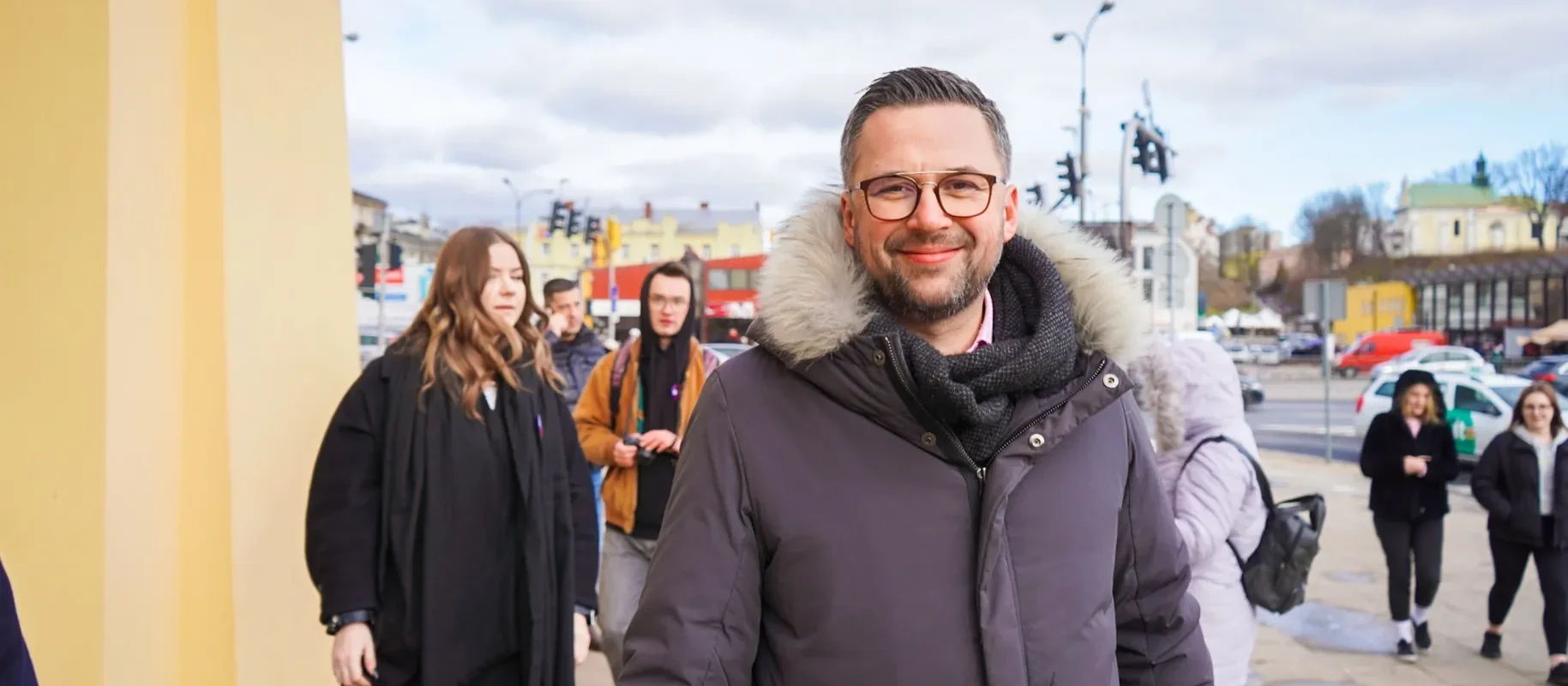 Na zdjęciu widać mężczyznę w ciepłej kurtce i okularach, który uśmiecha się, idąc w towarzystwie innych osób, na tle miejskiego krajobrazu.