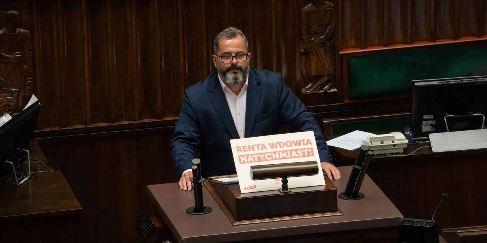 Mężczyzna w garniturze stoi za mównicą w sali obrad, a na biurku leży plakat z napisem „Renta wdowia natychmiast!”.