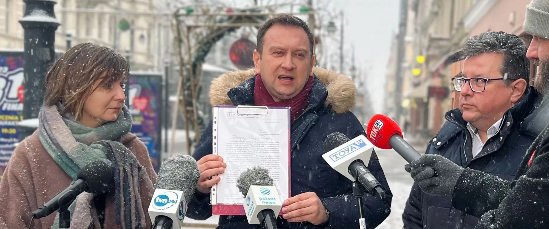 Mężczyzna w zimowej kurtce z futrzanym kapturem trzyma w dłoni dokument w teczce i zabiera głos do mikrofonów dziennikarzy na ulicy podczas zimowej konferencji prasowej.