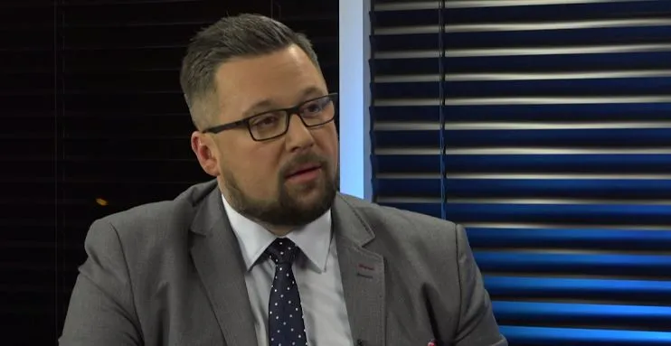 Mężczyzna w garniturze i okularach siedzi w studiu telewizyjnym, z czarnymi żaluzjami w tle.