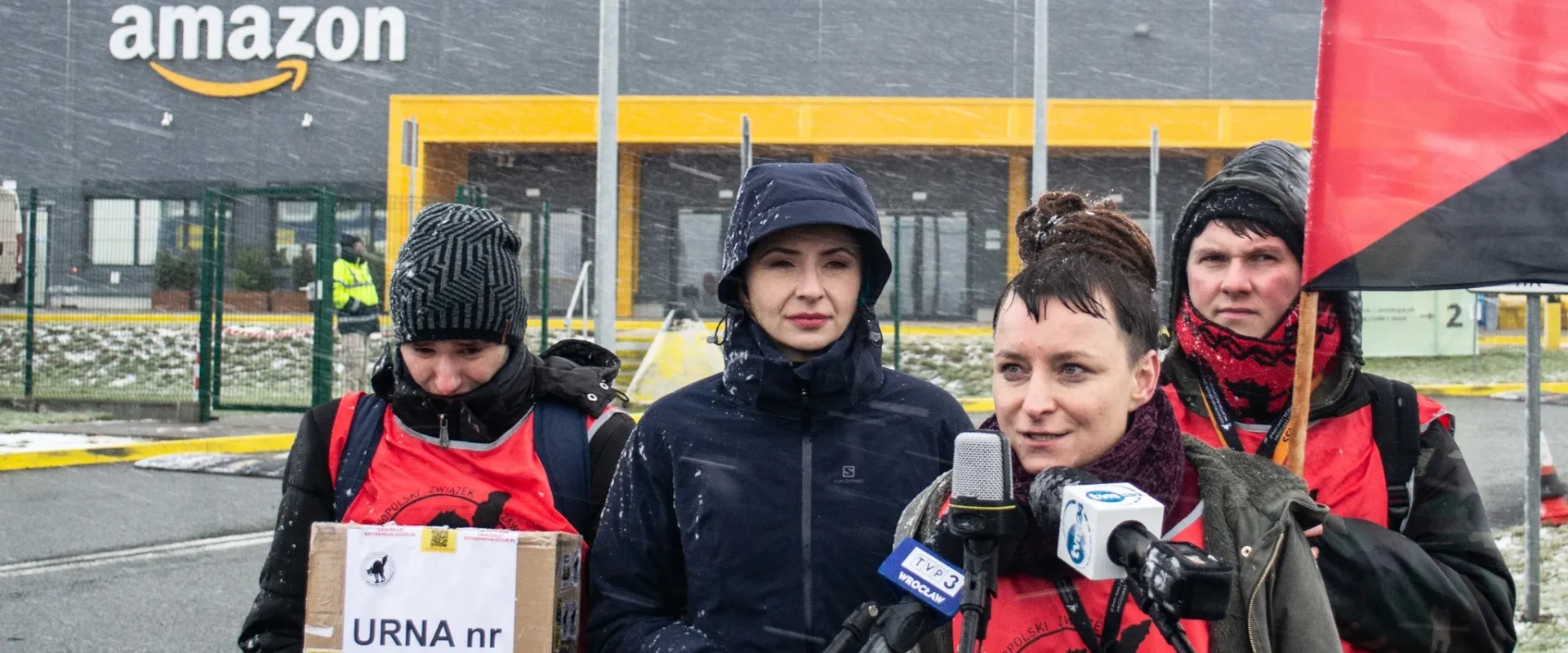 Grupa protestujących w czerwonych kamizelkach, z flagą, przemawia przed budynkiem Amazonu w śnieżnej scenerii.