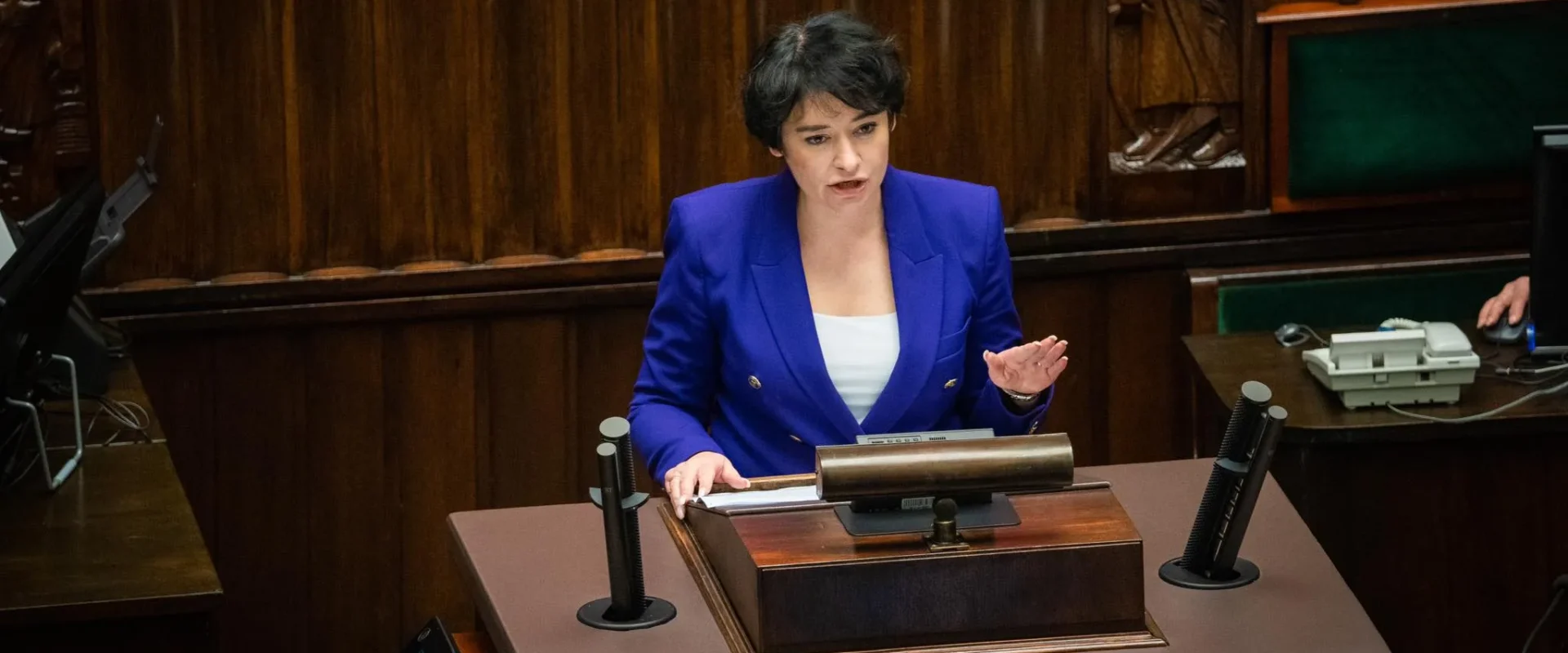 Kobieta w niebieskim garniturze przemawia z mównicy w sali posiedzeń z drewnianymi panelami w tle.