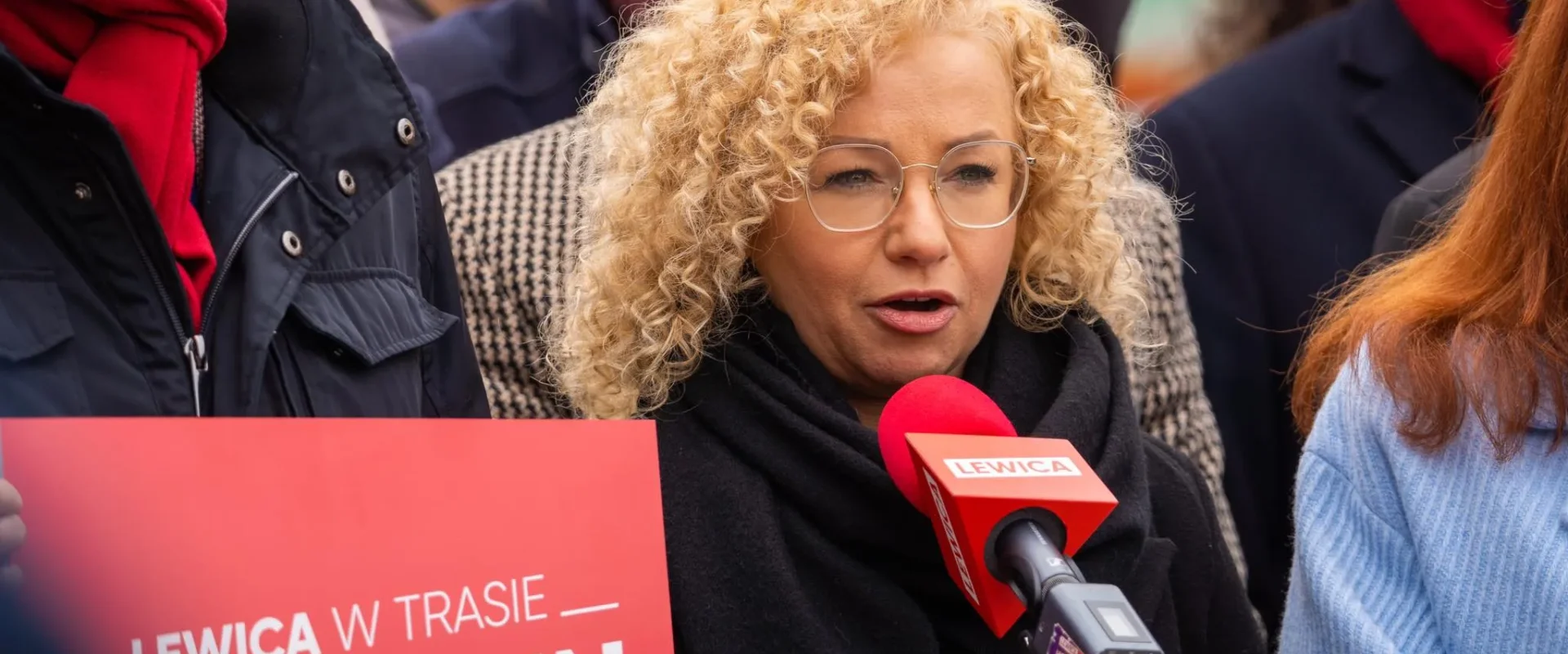 Kobieta z kręconymi blond włosami i okularami przemawia do mikrofonu z logo Lewica podczas demonstracji, stojąc obok czerwonego plakatu Lewica w Trasie.