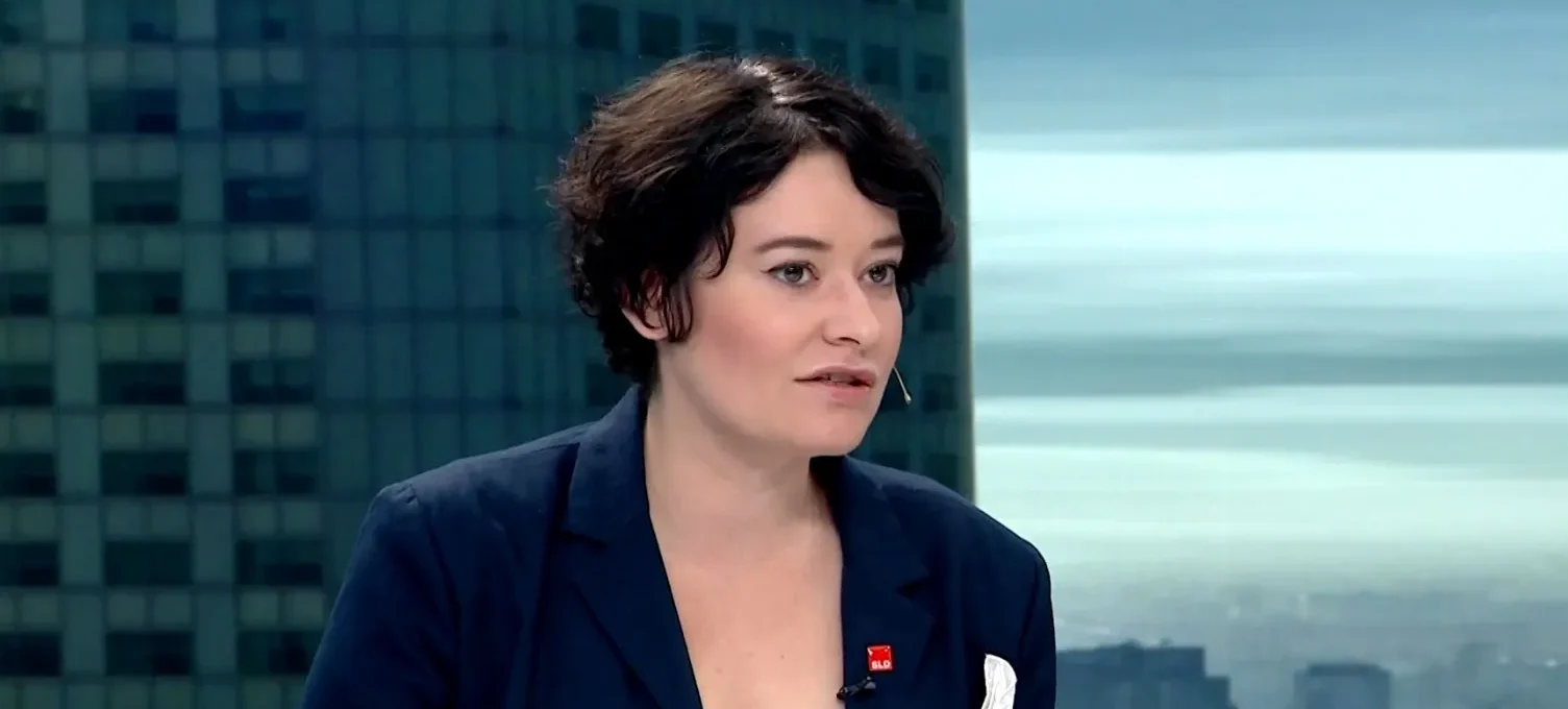 Kobieta o krótkich ciemnych włosach w granatowej marynarce rozmawia w studiu telewizyjnym, z mikrofonem lavalier przypiętym do ubrania, na tle panoramy miasta.
