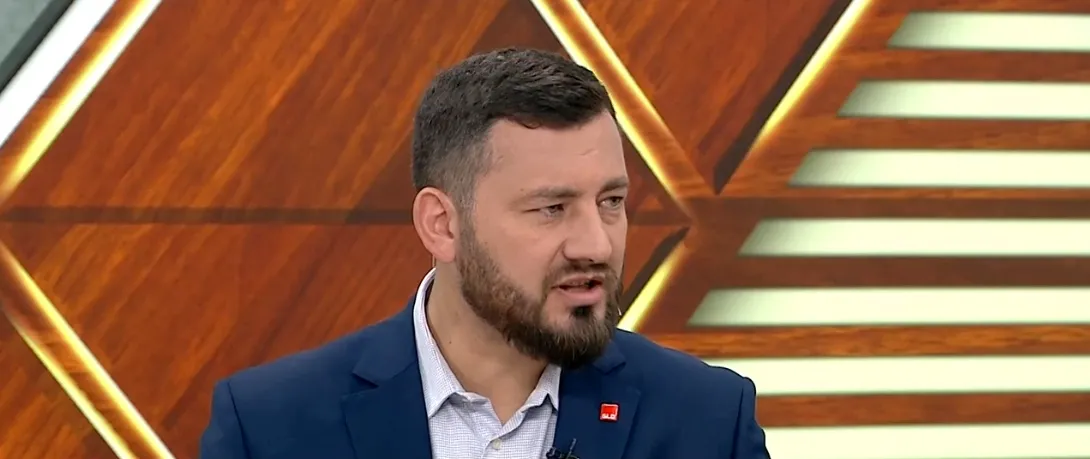 Mężczyzna z krótką brodą w granatowym garniturze i jasnej koszuli przemawia w studiu telewizyjnym, a w tle widać ozdobne drewniane elementy i żółte podświetlenie.