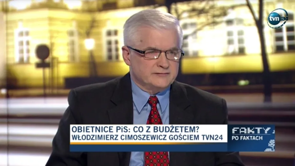 Mężczyzna w garniturze i okularach siedzi w studiu telewizyjnym podczas wywiadu, z dolnym paskiem informacyjnym na ekranie.