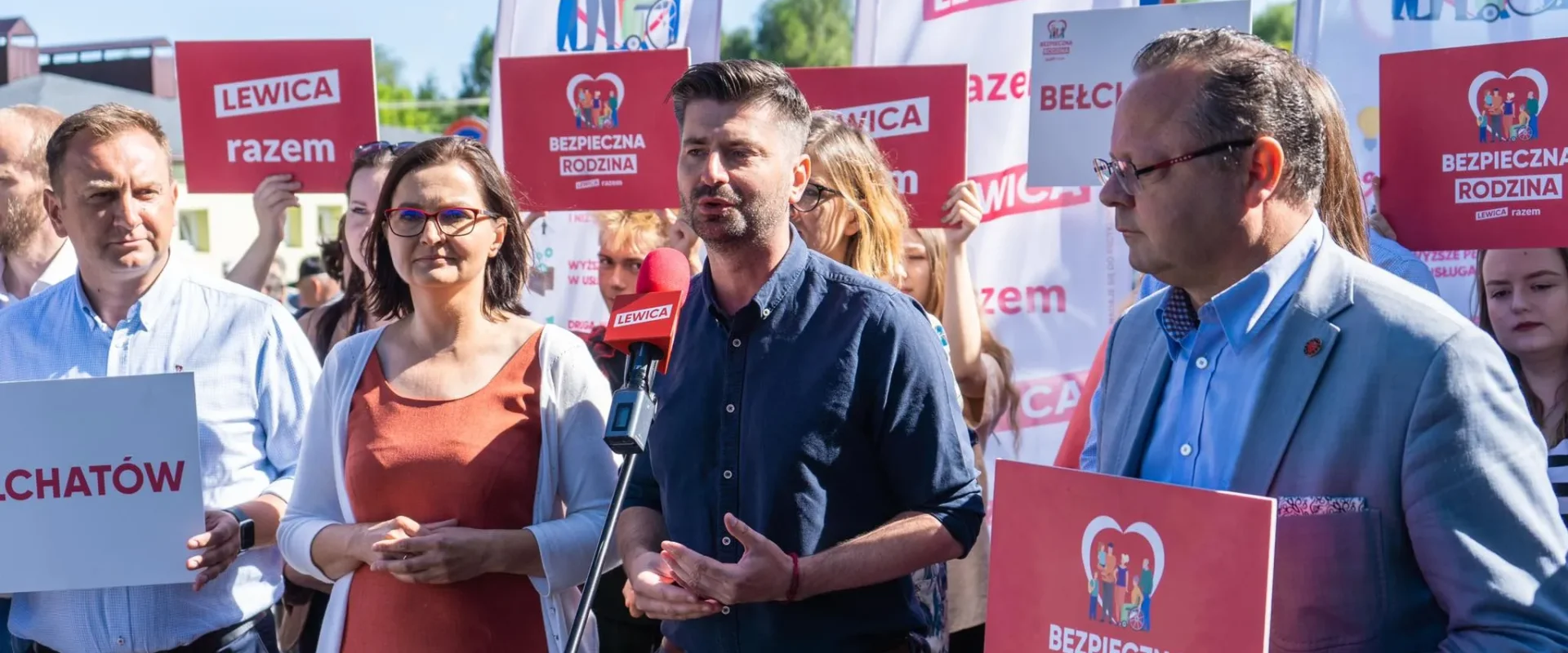 Grupa zwolenników partii Lewica na plenerowym wiecu; mężczyzna w granatowej koszuli przemawia do mikrofonu z czerwonym logo Lewica, a w tle widoczne są plakaty z napisem „Bezpieczna Rodzina” i „Lewica razem”.