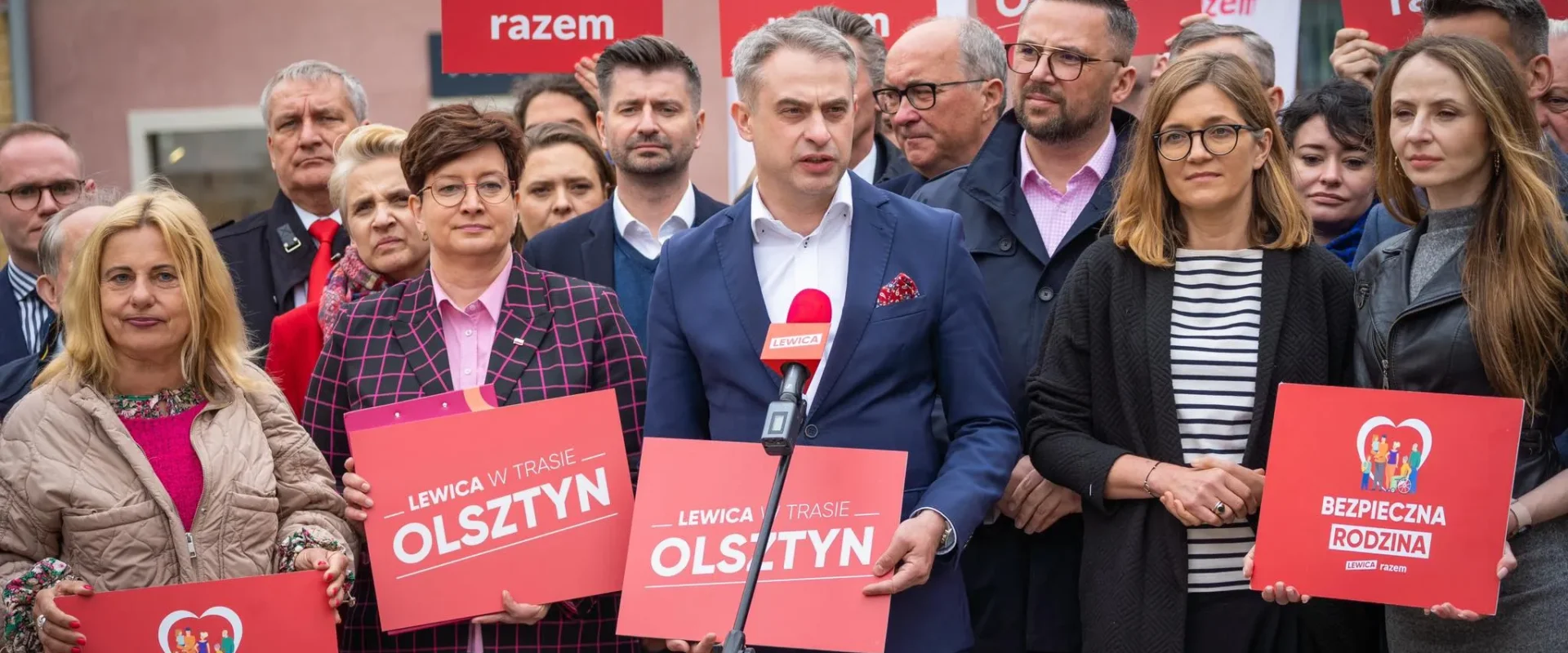 Grupa polityków Lewicy podczas konferencji prasowej, na pierwszym planie mężczyzna w granatowym garniturze mówiący do mikrofonu, otoczony innymi uczestnikami i czerwonymi plakatami z hasłami Lewica.