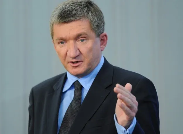 Mężczyzna w ciemnym garniturze i niebieskiej koszuli przemawia, gestykulując prawą ręką.
