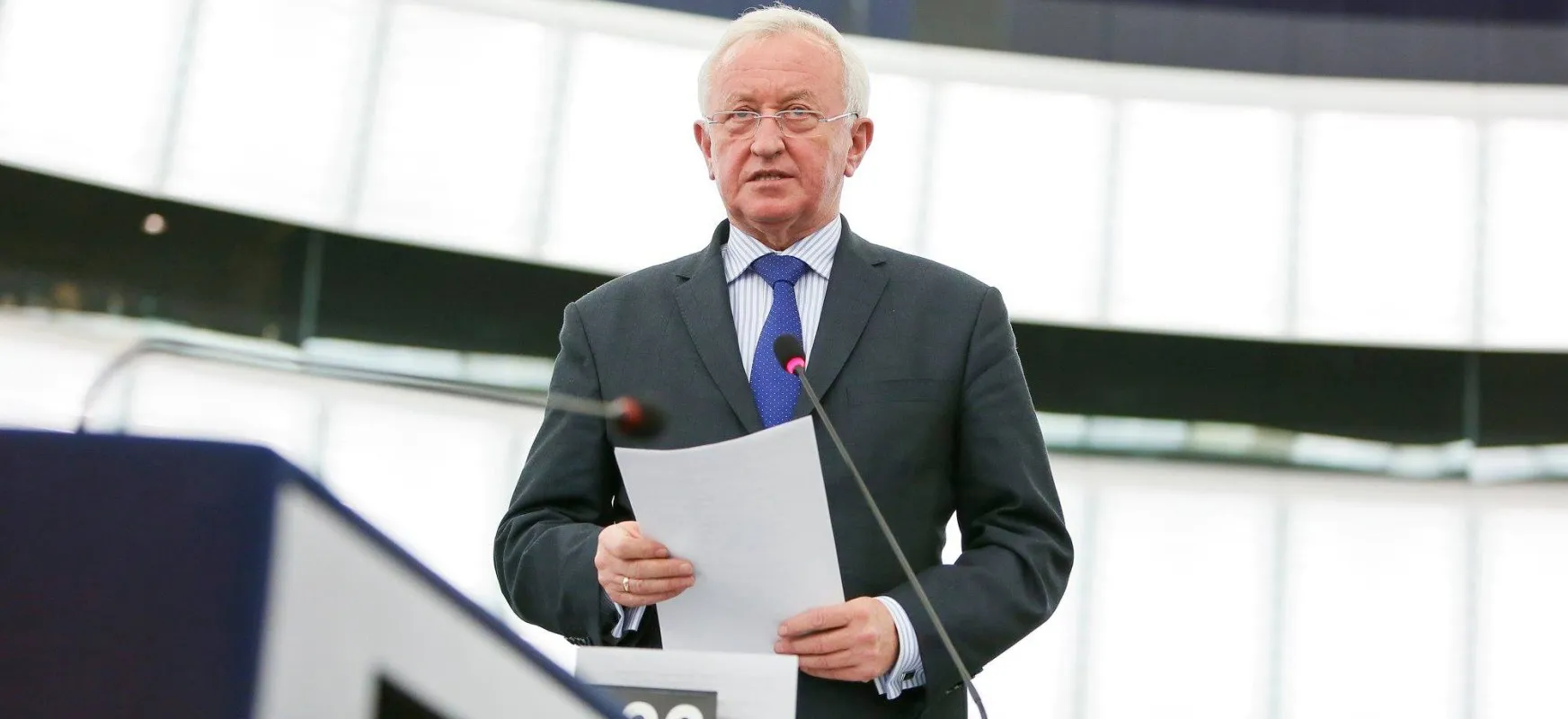 Mężczyzna w garniturze z kartkami w dłoni przemawia do mikrofonu podczas konferencji.