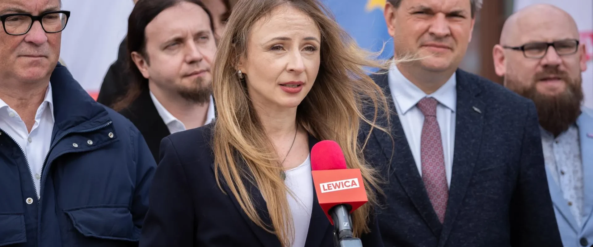 Kobieta przemawia do mikrofonu z logiem Lewicy podczas konferencji prasowej, otoczona przez grupę mężczyzn.