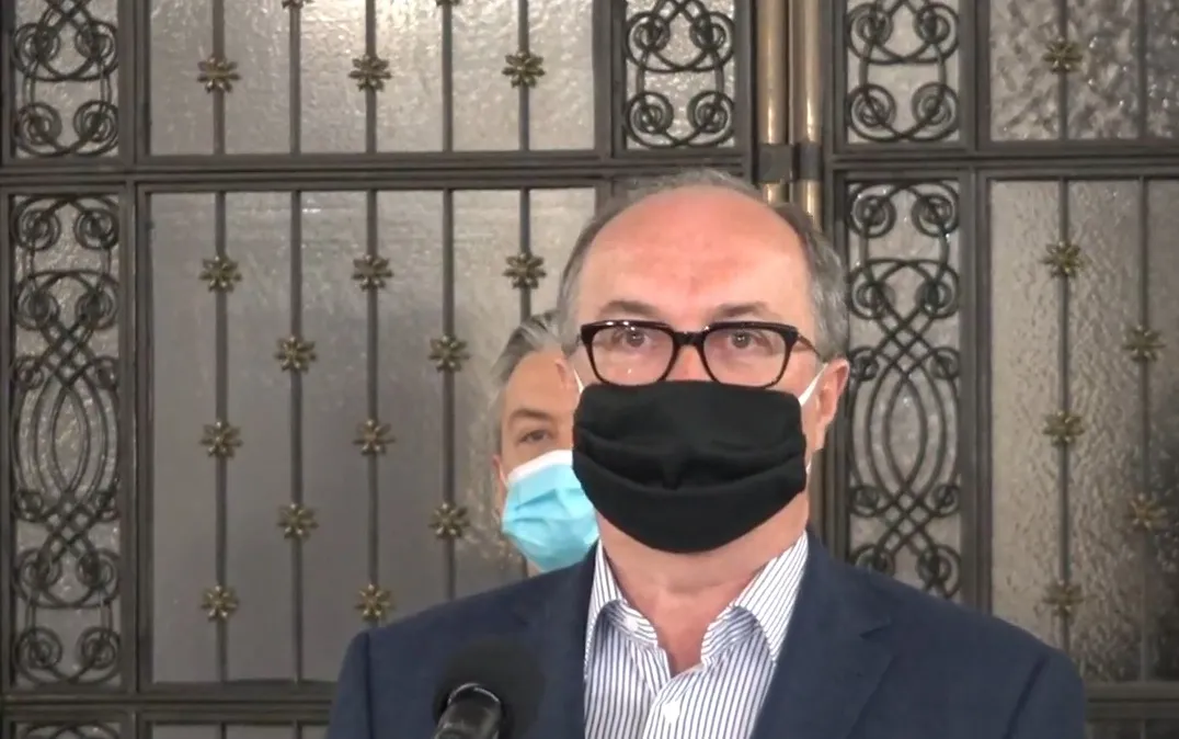 Mężczyzna w okularach, czarnym maseczce i garniturze stoi przed mikrofonem i przemawia do kamery, a w tle widoczna jest ozdobna metalowa brama oraz osoba w niebieskiej masce.