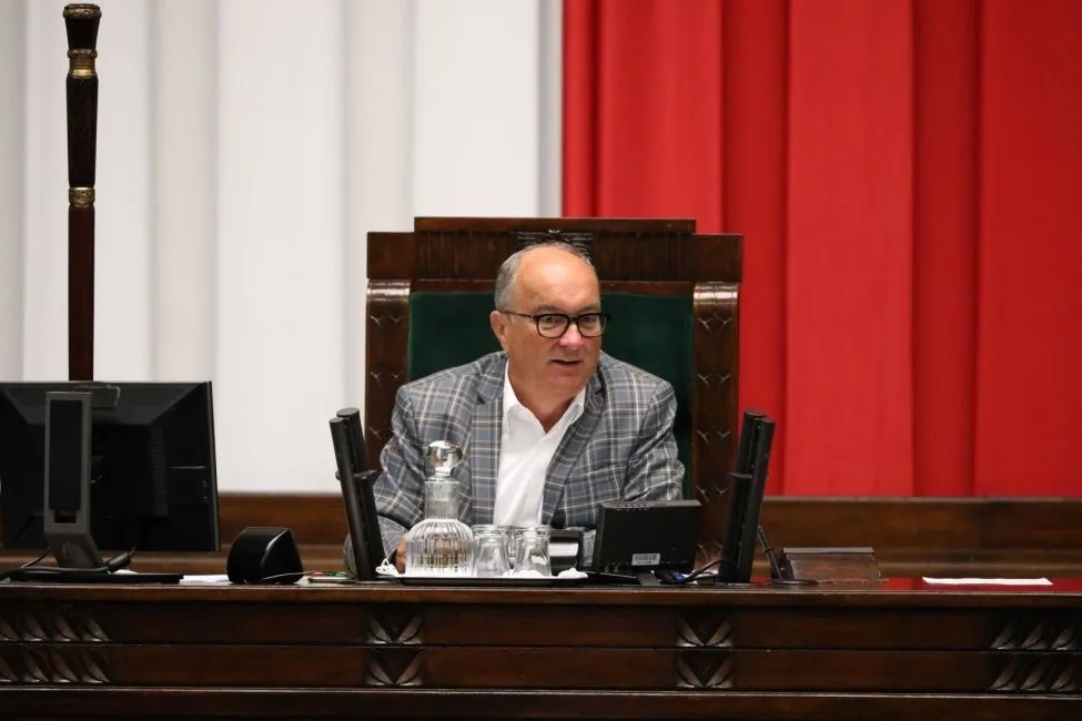 Mężczyzna w marynarce w kratę siedzi przy mównicy w sali obrad, na tle czerwonej zasłony, z monitorami i dzbanuszkiem z wodą na blacie.