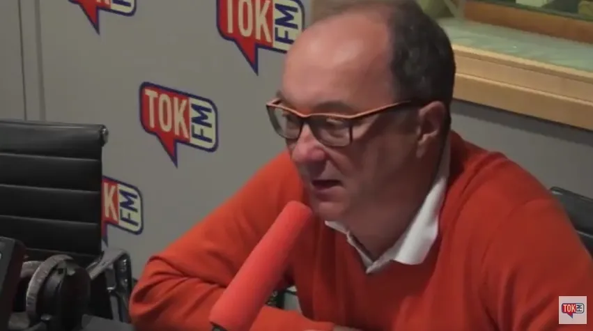 Mężczyzna w pomarańczowym swetrze i okularach mówi do mikrofonu w studiu radiowym z widocznym logiem TOK FM.