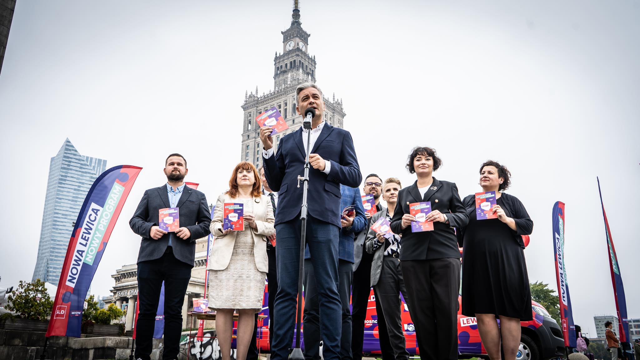 Kamper Nowej Lewicy na plenerowym wydarzeniu w Polsce; grupa działaczy stoi na scenie, lider przemawia do mikrofonu i trzyma ulotkę, w tle widoczne są banery i pojazd kampanijny z logo partii.