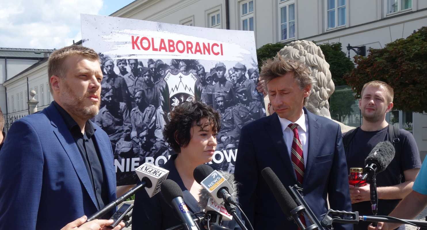 Grupa osób podczas konferencji prasowej stoi przed dużym plakatem z napisem „KOLABORANCI” i zdjęciem żołnierzy, a kobieta przemawia do mikrofonów.