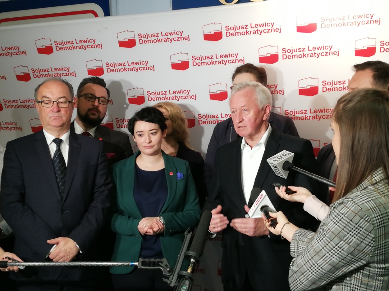 Grupa polityków Sojuszu Lewicy Demokratycznej podczas konferencji prasowej, stoją przed banerem z logotypami partii i odpowiadają na pytania mediów.
