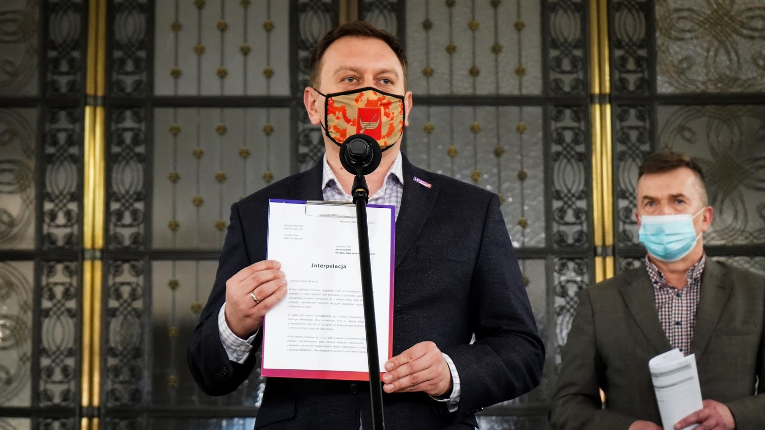 Mężczyzna w garniturze z maseczką trzyma dokument z napisem Interpelacja przy mikrofonie podczas konferencji; obok stoi drugi mężczyzna w maseczce, w tle ozdobne drzwi.