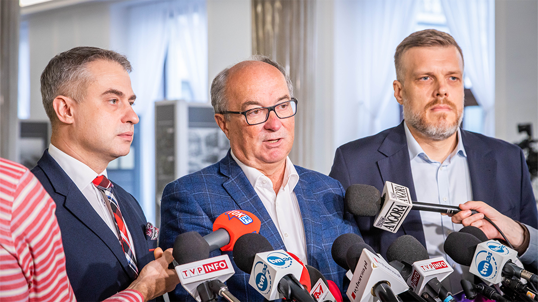 Na zdjęciu trzech mężczyzn stoi przed kamerami, a jeden z nich mówi do dziennikarzy, trzymając mikrofon.