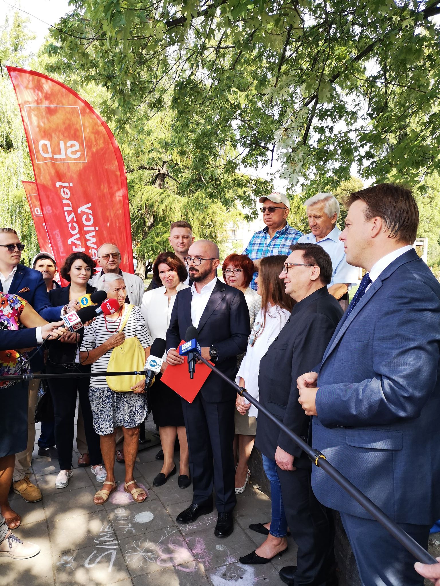 Liderzy listy do Sejmiku i Rady Warszawy komitetu SLD Lewica Razem przemawiają do mediów podczas konferencji prasowej, otoczeni dziennikarzami i współpracownikami przy czerwonych flagach partii.