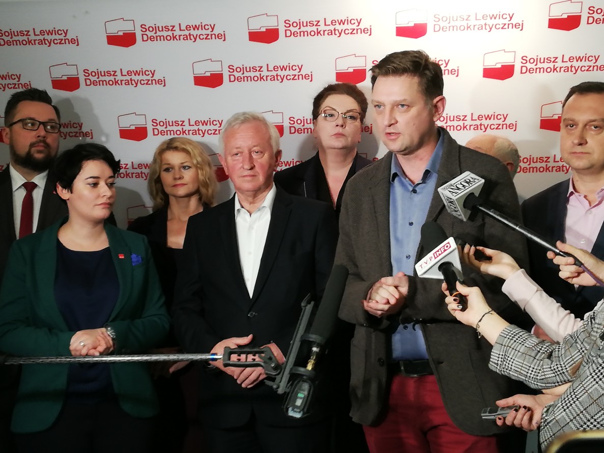 Grupa polityków Sojuszu Lewicy Demokratycznej podczas konferencji prasowej, mężczyzna po prawej przemawia do mikrofonów otoczony przez dziennikarzy, a w tle widnieje wielokrotnie logo partii.