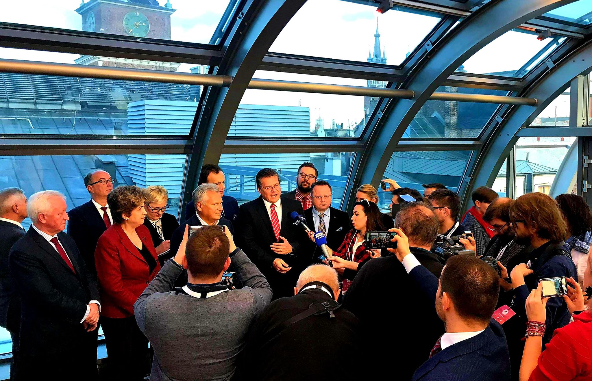 Grupa polityków w garniturach udziela konferencji prasowej w szklanym, łukowatym atrium, otoczeni przez reporterów i kamery.