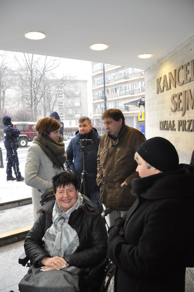 Grupa osób przed wejściem do Kancelarii Sejmu w zimowej scenerii, kobieta na wózku inwalidzkim uśmiecha się do aparatu.