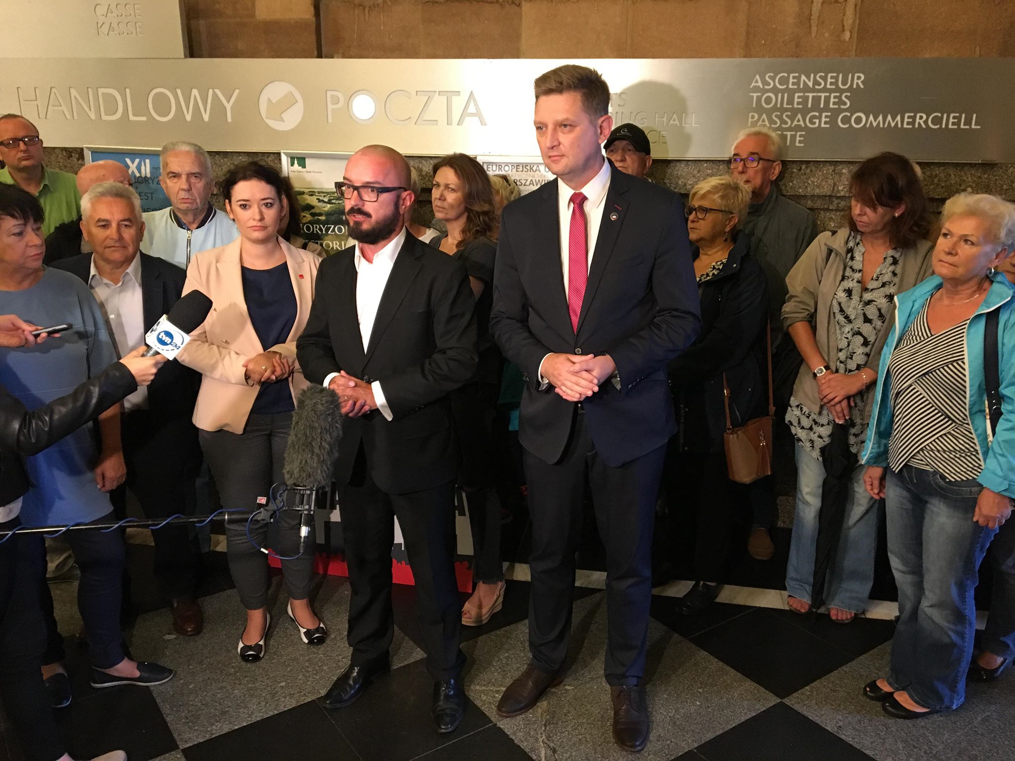 Grupa osób zgromadzona w holu podczas konferencji prasowej; dwaj mężczyźni w garniturach na pierwszym planie prowadzą briefing, otoczeni przez reporterów i obserwatorów.