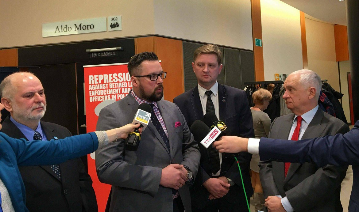 Kilku mężczyzn w garniturach udziela oświadczenia przed kamerami i mikrofonami na konferencji prasowej, obok banera o represjach, w kontekście dyskusji o odpowiedzialności zbiorowej w Polsce.